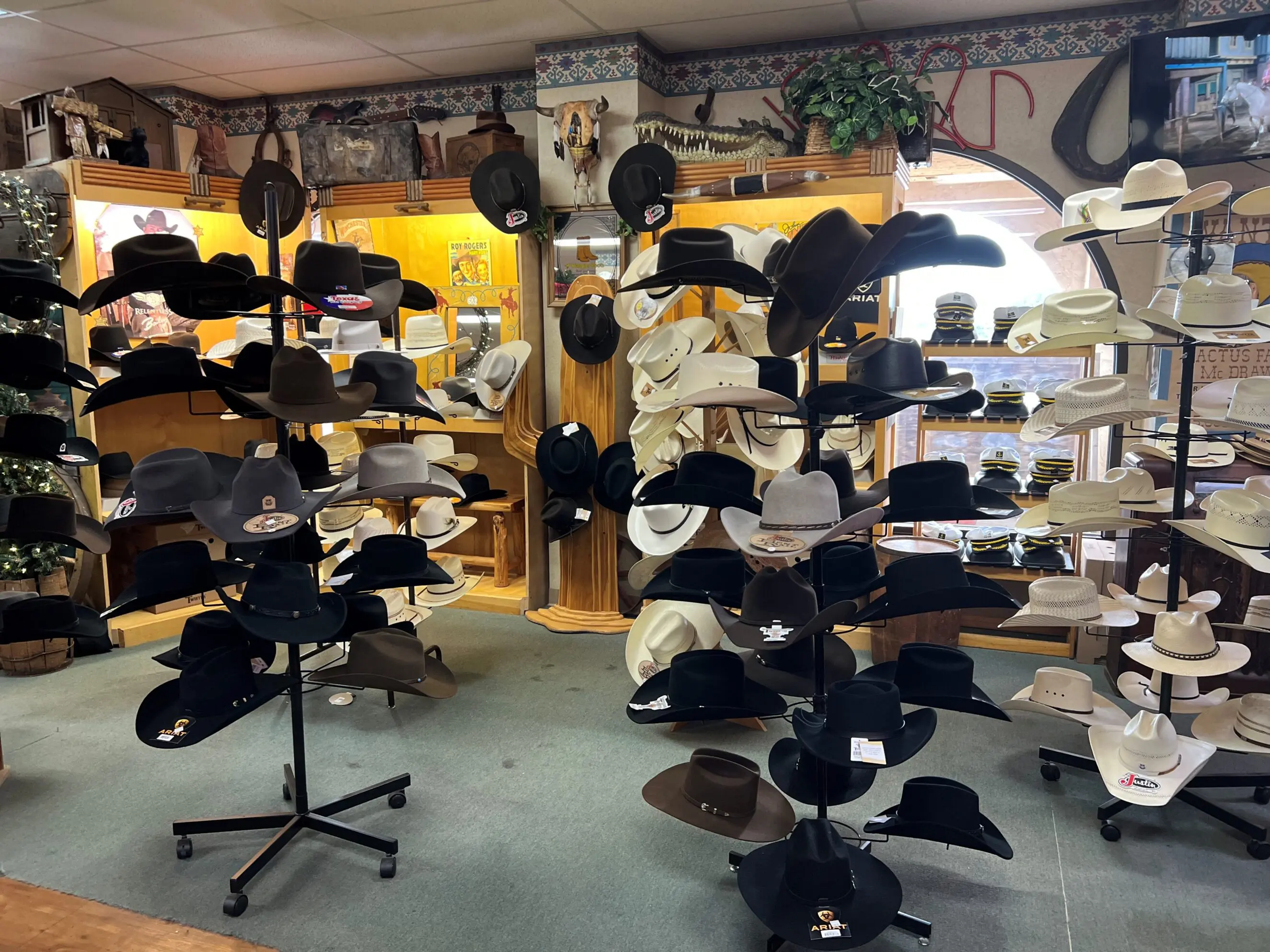 Inside a family‑owned western wear shop in Cusseta, Alabama: multiple rotating hat racks filled with black, brown, gray and white cowboy hats and straw hats arranged on stands and shelves against wood display cabinets and a decorated wall, showcasing a variety of ranch and dress styles for boots, workwear and everyday western fashion