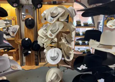 Display stand of cream and white cowboy hats with black and gray hats on surrounding racks inside a western hat shop, shelves of folded caps and hat price tags visible in the background