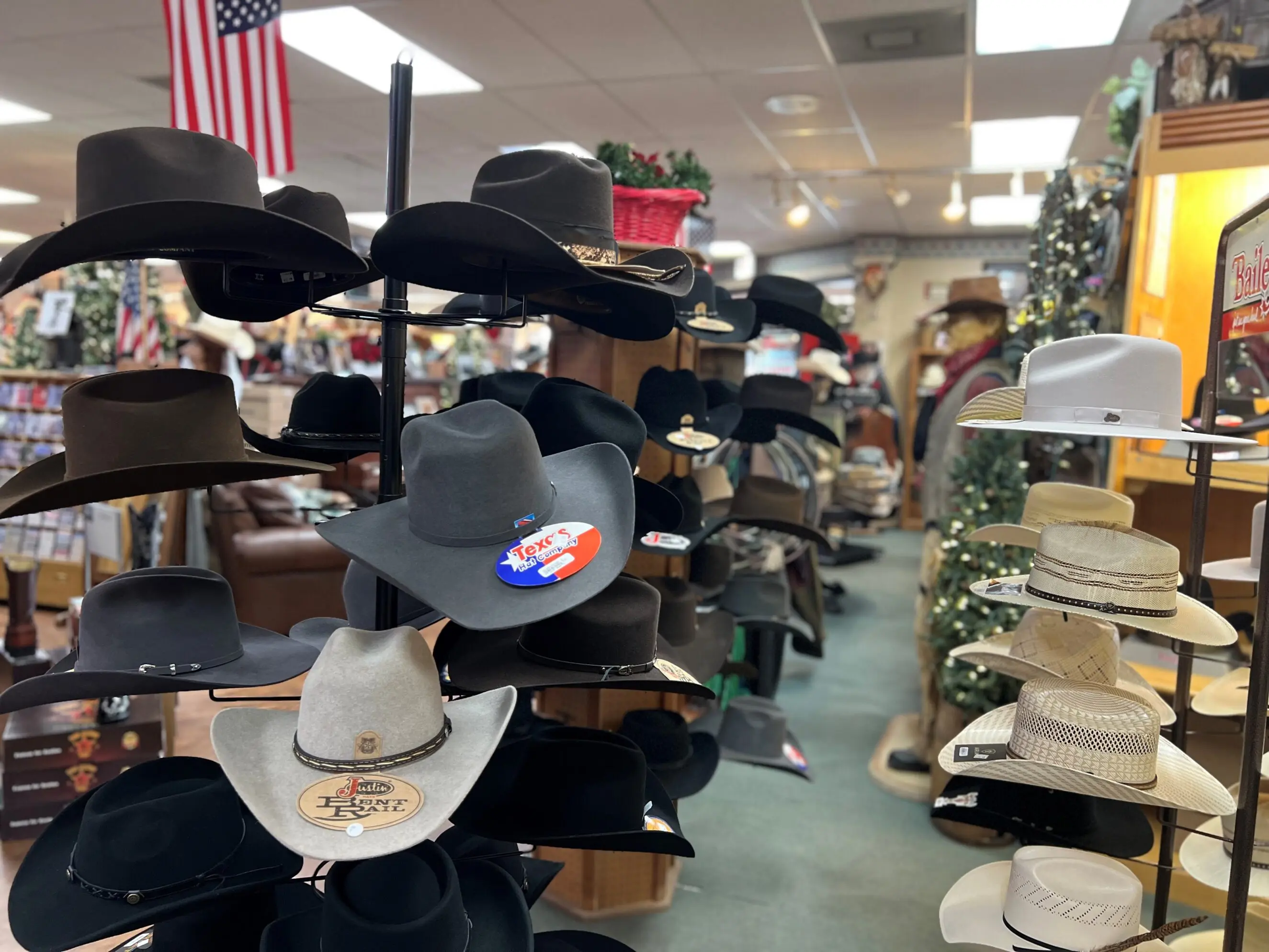 Display of assorted western hats on rotating racks in a country store aisle, featuring felt and straw cowboy hats in black, gray, tan and white with brand stickers, surrounded by shelves of western apparel and an American flag overhead