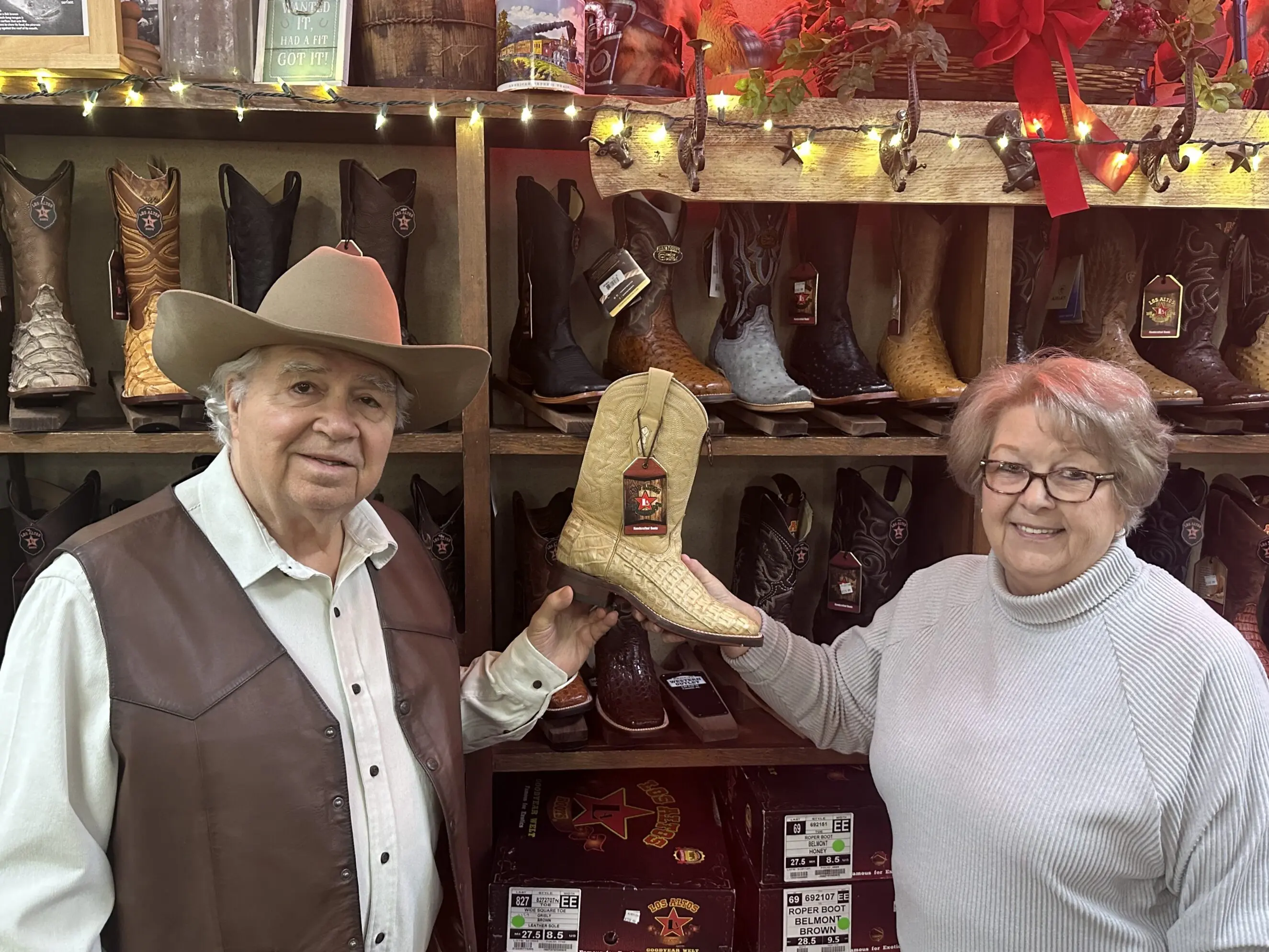 Two people in a western wear store holding up a tan crocodile-pattern cowboy boot in front of wooden shelves filled with assorted cowboy boots, boot boxes, string lights and rustic decor, evoking a family‑owned local boot shop.