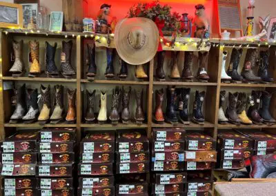 Rows of men's western boots in various leathers and colors displayed on wooden cubed shelves with a straw cowboy hat hanging center, string lights and decorative items above, and stacked shoeboxes below in a country store boots section