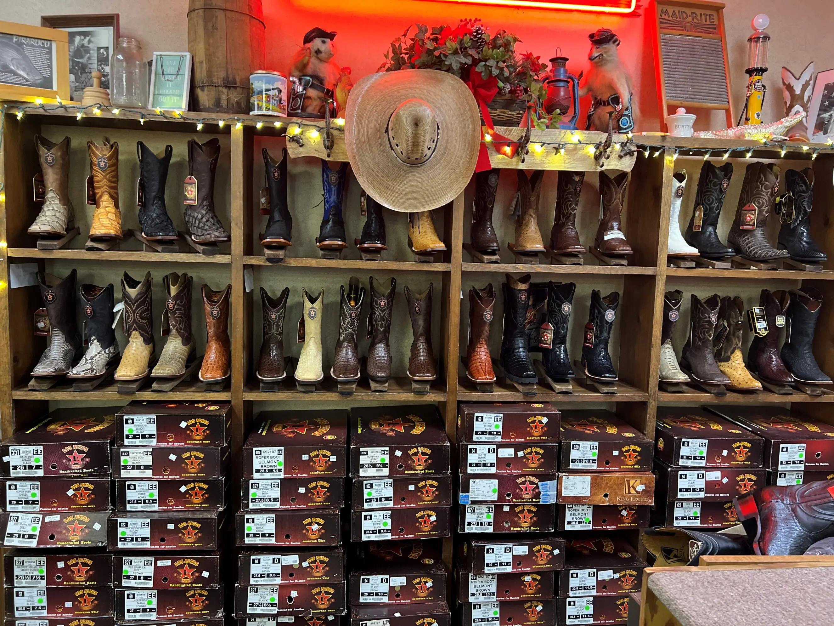 Shelves of western boots in a family-owned Bridges Western Wear shop: multiple rows of men’s and women’s leather cowboy boots in various colors and textures displayed on wooden cubbies with stacked shoeboxes below, a straw cowboy hat hanging center, festive string lights and Western decor on the top shelf reflecting the Auburn/Opelika-area western wear store atmosphere