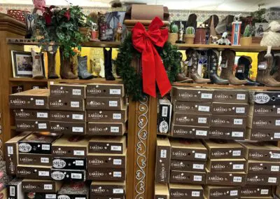 Stacked Laredo boot boxes filling wooden shelving in a western store display, with cowboy boots and decorative items on the top shelves and a green wreath with large red bow hung on a carved wooden center post.