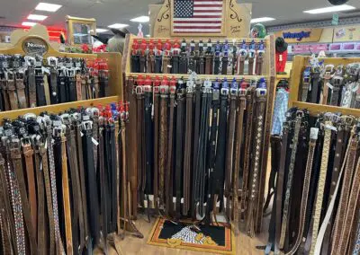 Wide display of leather belts in a western store: multiple wooden racks filled with brown, black, embossed and woven belts with metal buckles, an American flag sign above, Wrangler signage and western shirts visible in the background, and a small eagle rug on the wooden floor.