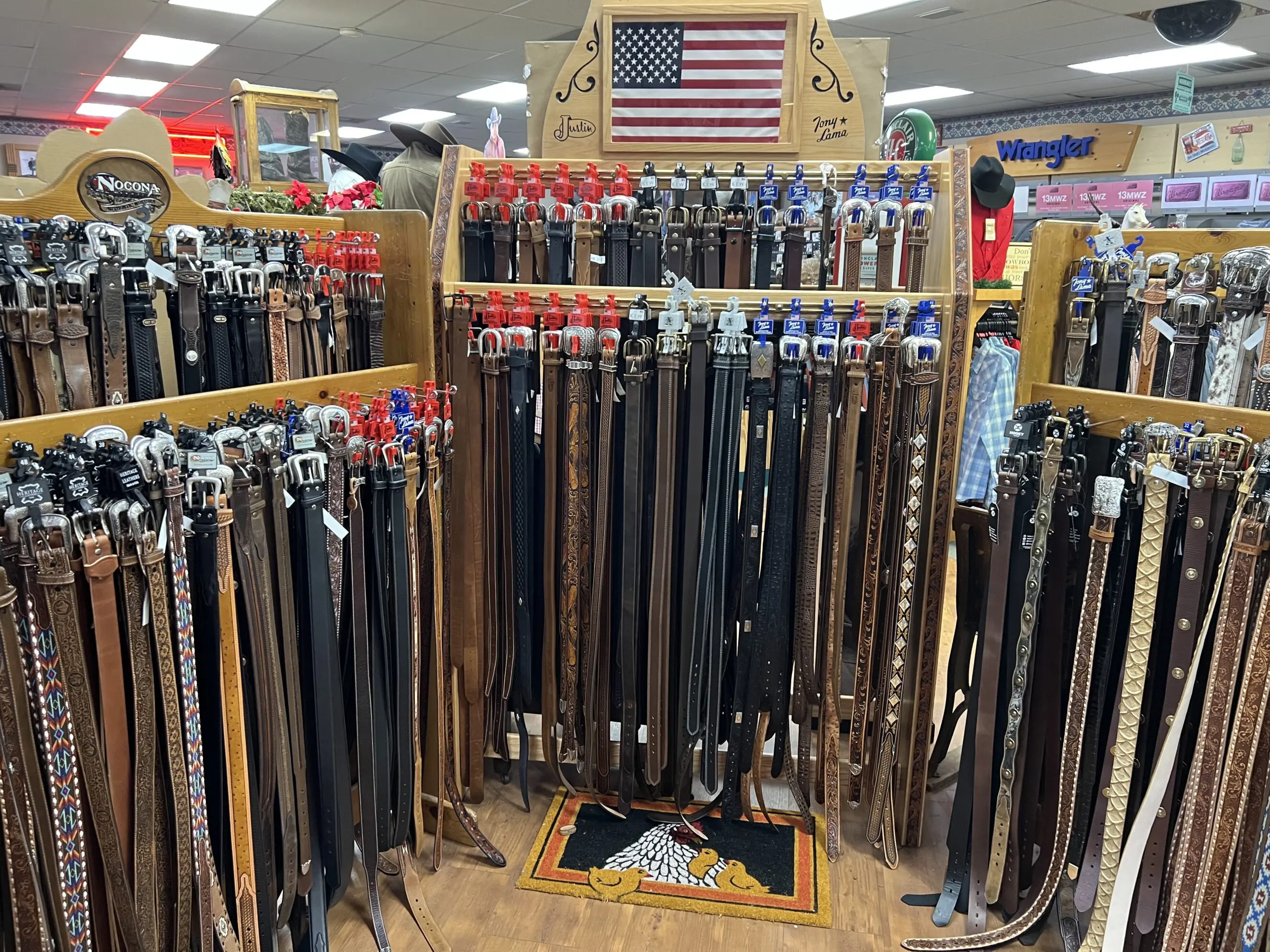 Display of dozens of western leather belts and decorative buckles hanging on wooden racks in a western wear store, with patterned and plain brown and black belts, branded tags visible, a small American flag mounted above, and Wrangler signage in the background