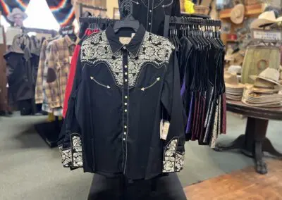 Black western-style long-sleeve button-up shirt with white floral embroidery on the yoke and cuffs, gold arrow pocket accents and snap buttons, hanging on a rack in a rustic clothing store with hats and other garments in the background
