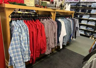 Display of men's western shirts on hangers in a retail store: light blue plaid shirts, bright red shirts, red-check shirts and assorted denim and solid shirts on a wooden rack with folded jeans on shelves in the background