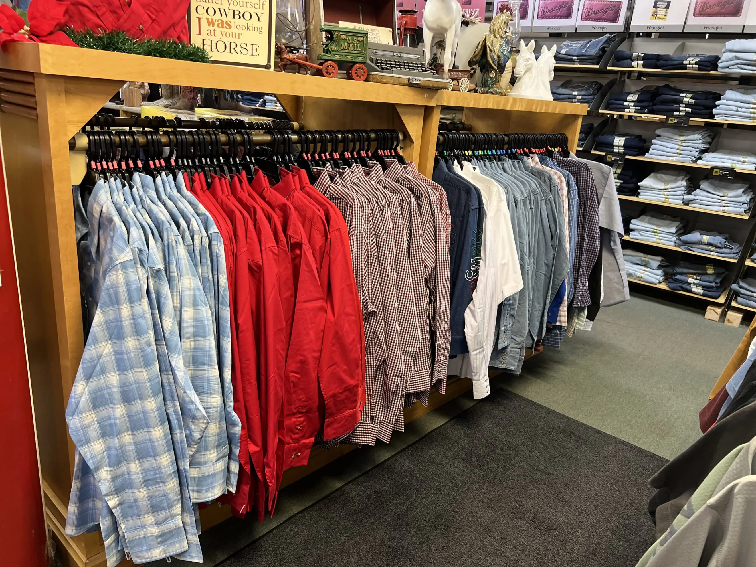 Row of men's western shirts in a retail display—light blue plaids, bright red solids, checkered and denim shirts hanging on wooden racks with folded jeans on shelves nearby, showing western wear apparel selection