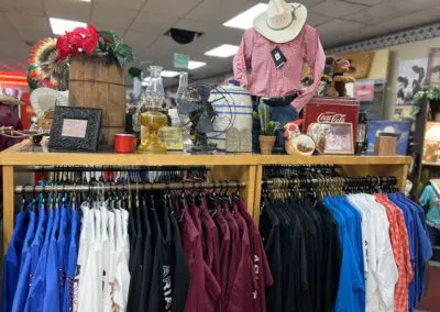 Mannequin wearing a white cowboy hat and red check shirt stands behind a wooden display table topped with rustic decor—wooden barrel, oil lamps, vintage fan, Coca‑Cola crate, ceramic rooster and small potted cactus—with two lower racks of neatly hung western shirts in blue, white, black and maroon inside Bridges Western & General Store.
