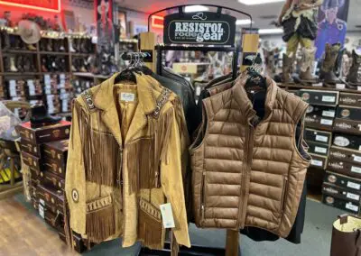 Western store display of tan suede fringe jacket with decorative stitching next to a quilted brown puffer vest on a Resistol rodeo gear rack, surrounded by boot boxes and cowboy boots in the background