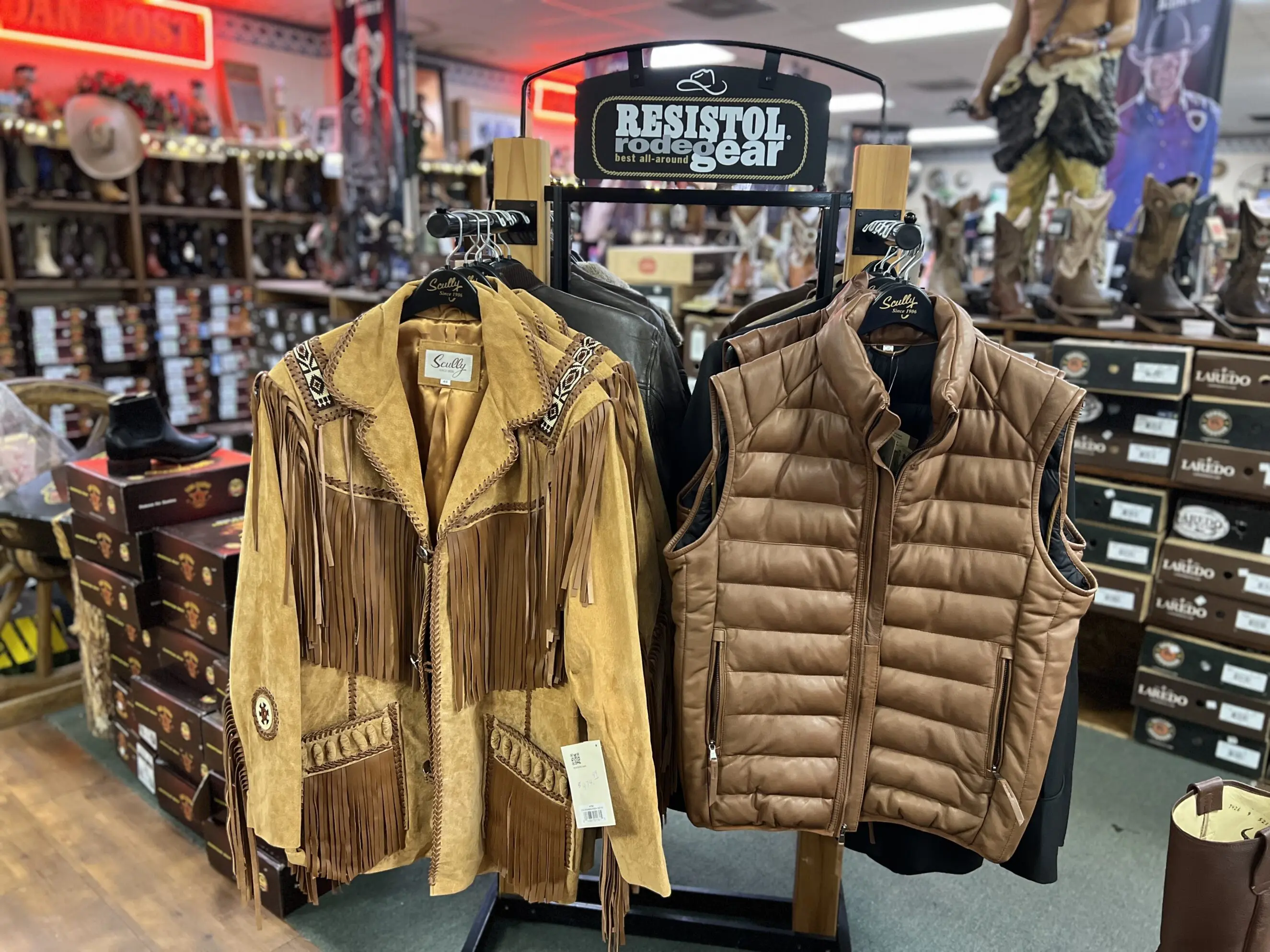 Suede fringed western jacket with bead and braid accents displayed next to a quilted brown puffer vest on a rack in a western wear store, with boot boxes and shelves of cowboy boots visible in the background
