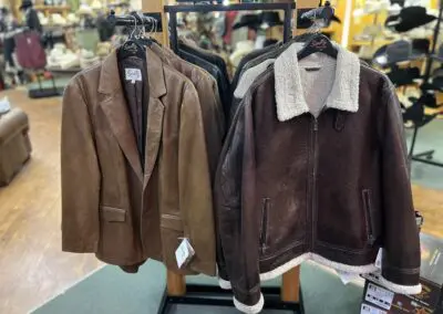 Two brown leather jackets on hangers in a retail store: a lighter brown tailored blazer-style leather jacket on the left and a darker brown shearling-collar zip-up leather jacket with cream fleece trim on the right, displayed on a wooden rack with other clothing and store interior visible in the background