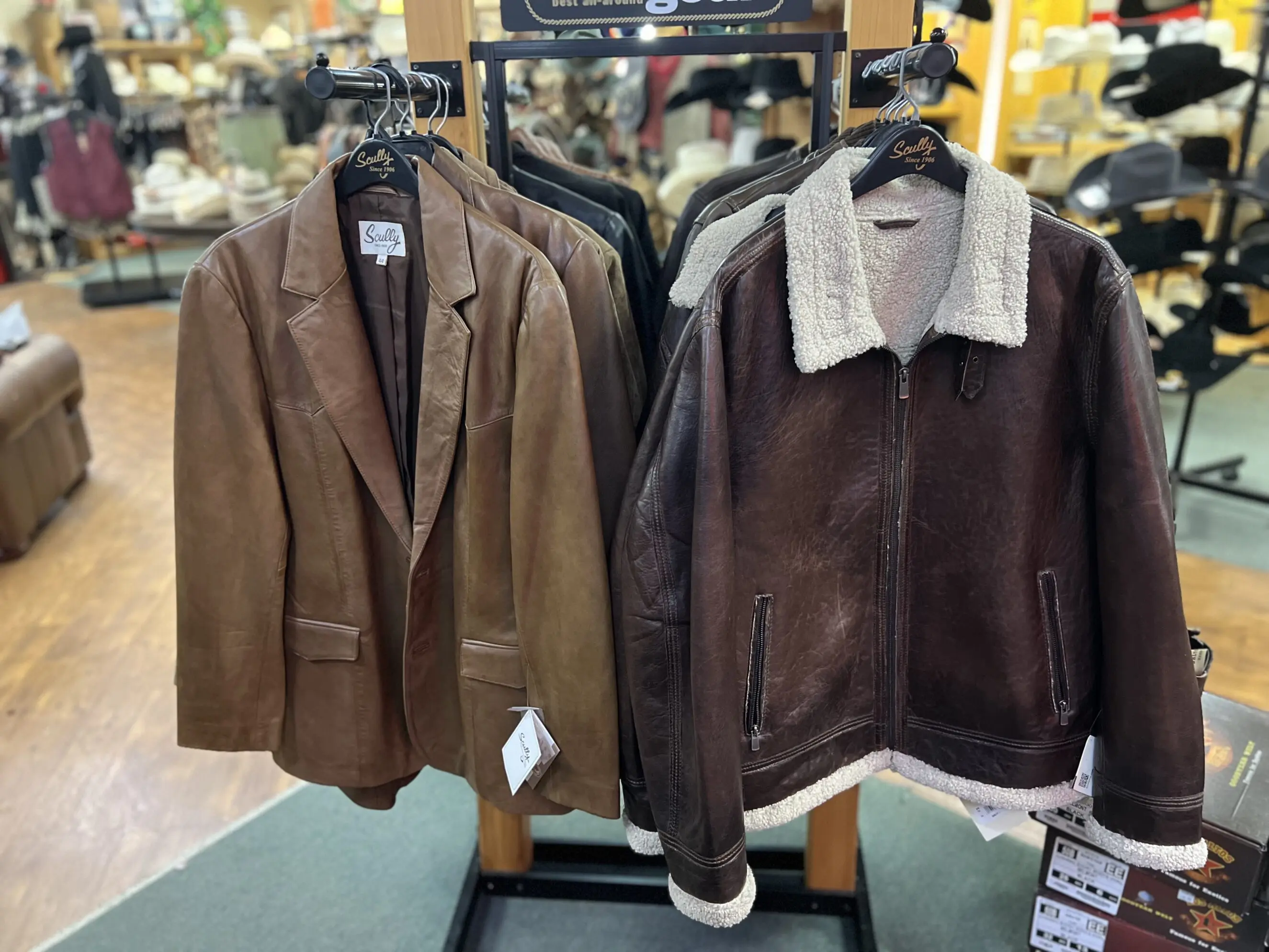 Two brown leather jackets on hangers at a western wear store rack — a tan leather blazer and a dark brown shearling‑collar bomber jacket displayed among other jackets with store floor and hat displays visible in the background