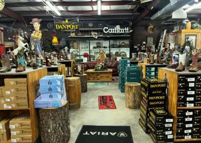 Wide interior view of a rustic western boot store showing wooden display tables, stacked Dan Post and Ariat shoe boxes, multiple cowboy boots arranged on pedestals, barrel and log stump props, branded signs (Dan Post, Carhartt) on the back wall, western decor including a large rooster statue and wagon wheels, rugs with store logos on concrete floor, and shelving with leather furniture and decor in the background.