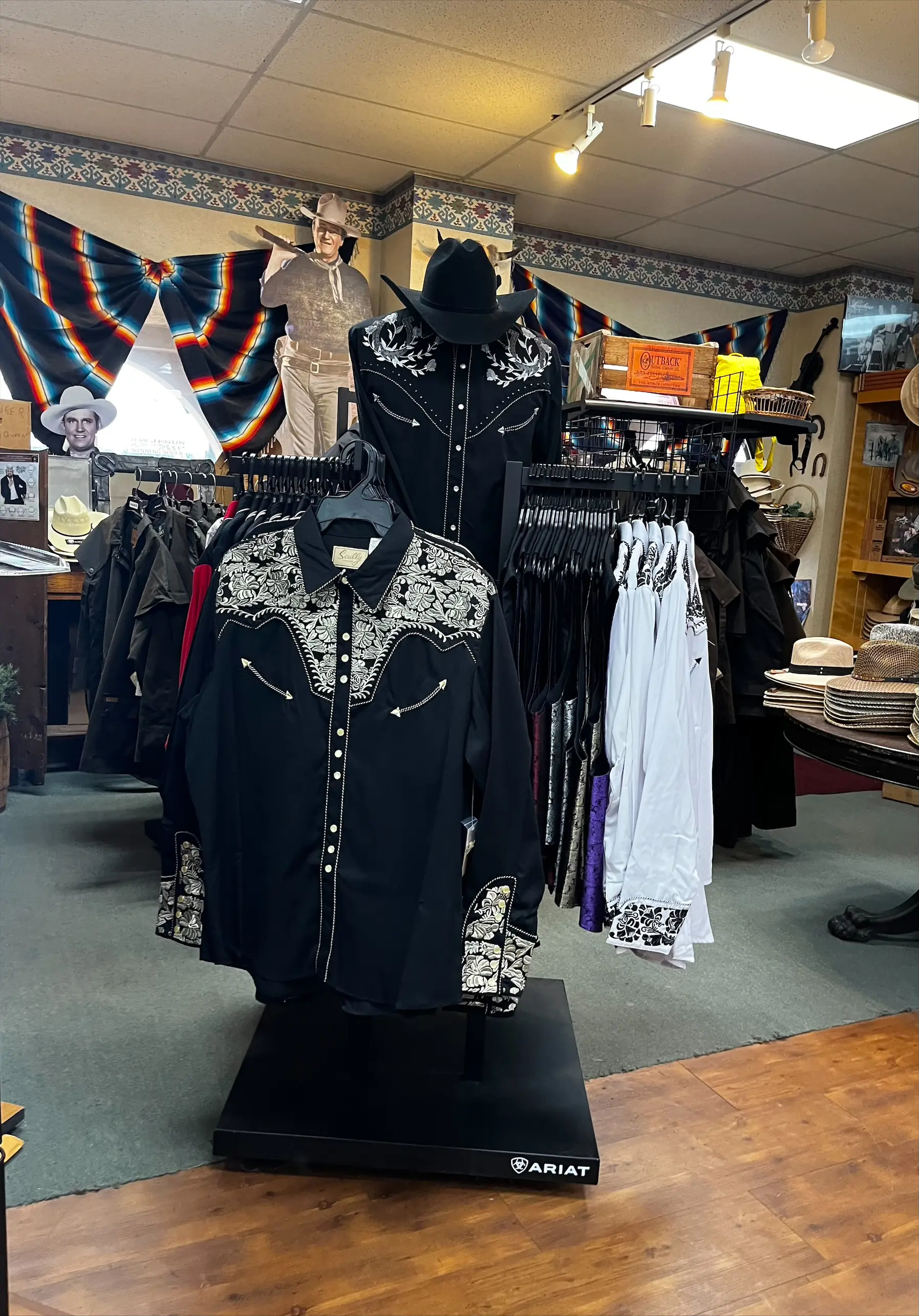Mannequin display of black western shirts with white floral embroidery and snap buttons on an Ariat floor stand, surrounded by racks of hats, shirts, and western accessories in a boutique store interior.